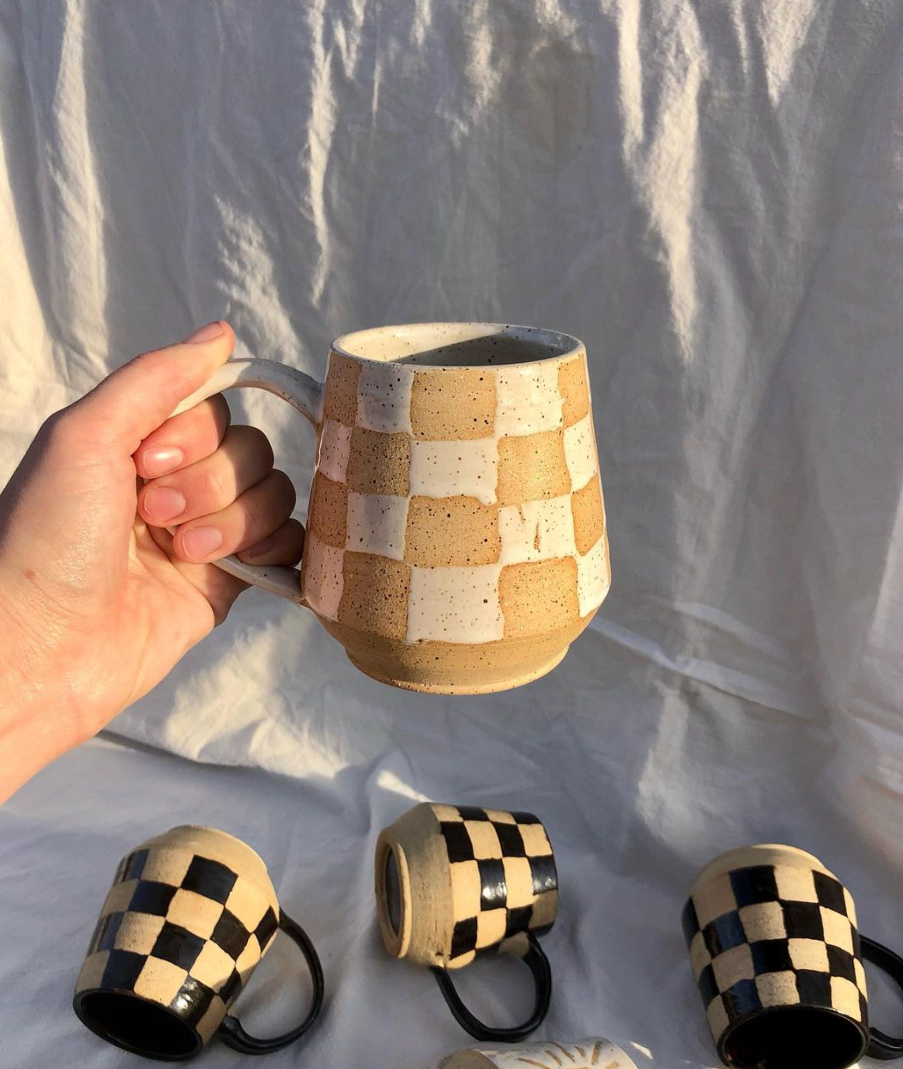 Person holds checker print ceramic mug in white + tan in front of white + black ceramic mugs, created by Cora Freeman Design