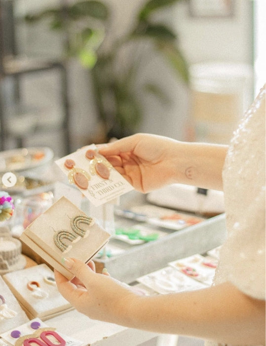 person holding up two pairs of earrings at the accessories table at Darling x Dashing